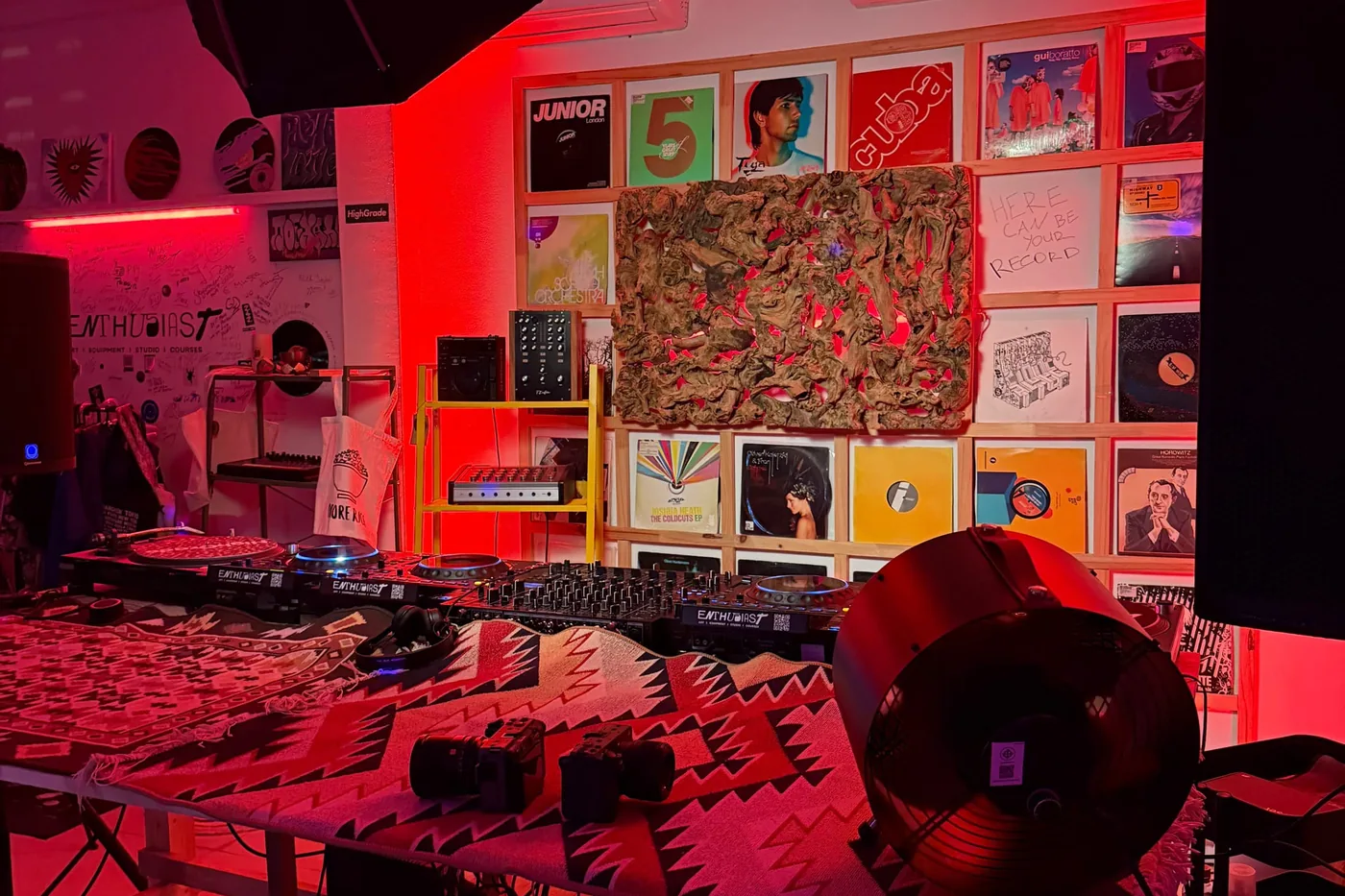The Enthusiast Place studio lit deep red, a grid of framed record sleeves covering the back wall behind a CDJ and mixer setup, a patterned rug running across the front of the booth.