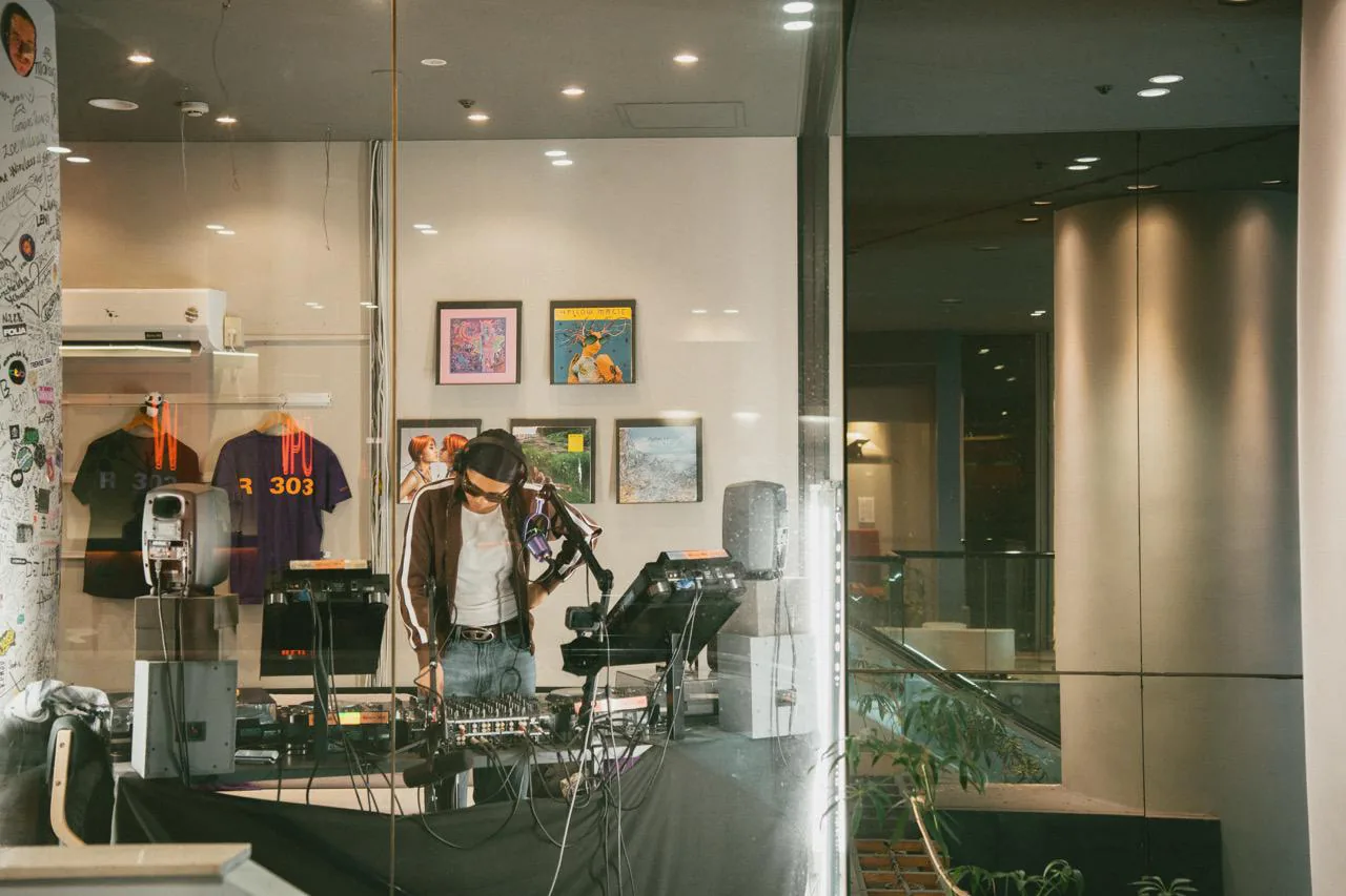 A DJ on air behind the glass frontage of the Room 303 studio, mixer and monitors in front of them, R 303 shirts and record sleeves on the wall behind.