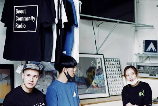 Three people standing in the studio in front of a rail of hanging Seoul Community Radio t-shirts, framed artwork and a rack of equipment along the wall behind them.