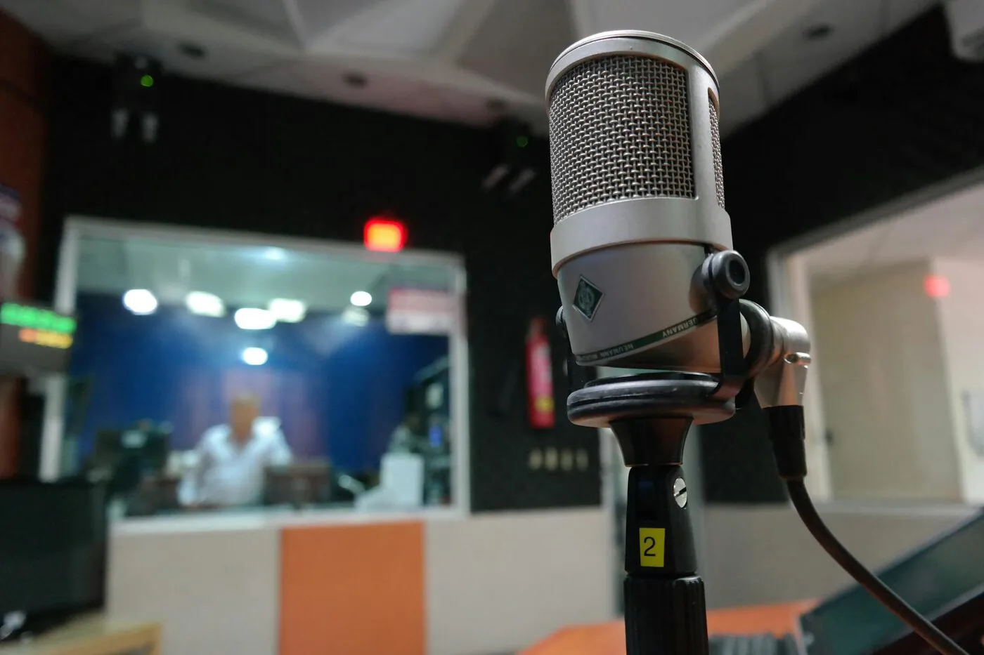 Close-up of a microphone in a recording studio with a blurred sound engineer behind glass.